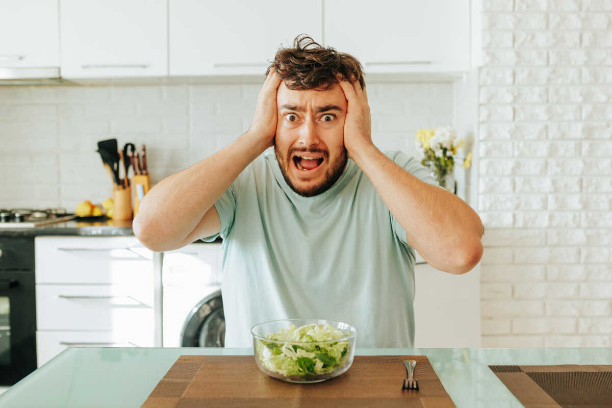 Sitting in kitchen and hold head with his hands, a young man looks desperately into the camera. Tired of eating lettuce and leafy greens. Healthy food and favorite cuisine. Healthy food concept.